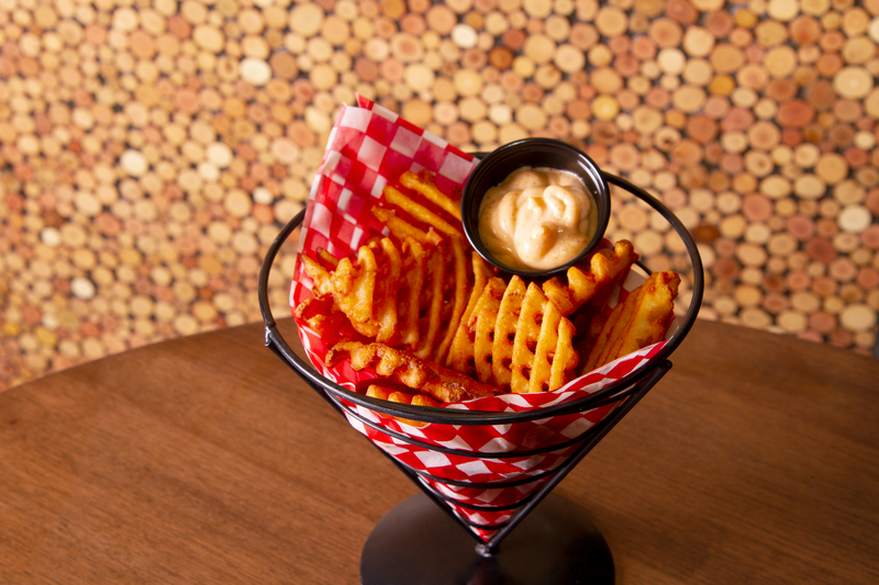 A cone-shaped wire basket with a red and white checkered paper liner, filled with seasoned crinkle-cut fries and a small round cup of dipping sauce, sitting on a wooden table with a cork-covered surface.