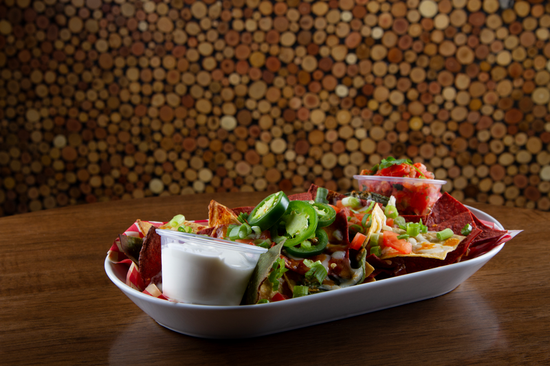 A boat-shaped dish of loaded nachos on a wooden table. The nachos are topped with melted cheese, diced tomatoes, shredded lettuce, and sliced jalapeños. Accompanied by sides of sour cream and salsa, the dish is set against a background featuring a cork-covered wall.