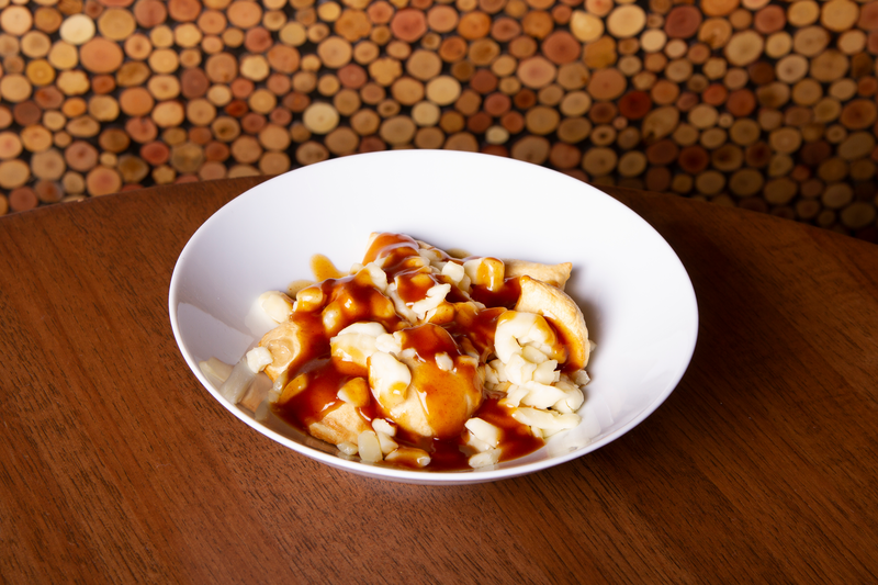 A bowl of poutine with pierogies on a wooden table. The dish includes cheese curds and a generous amount of brown gravy poured over the pierogies, presented in a white bowl. The setting has a background of a cork-covered wall.